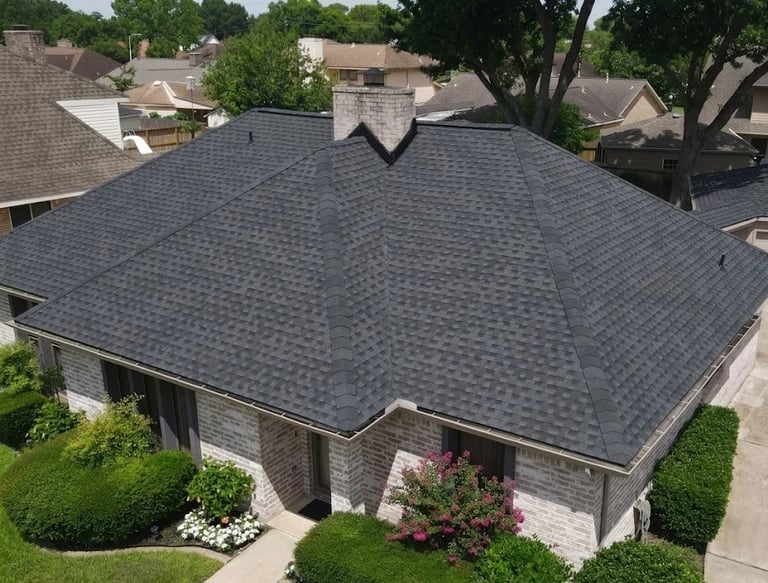 Aerial view of a modern residential house with dark gray asphalt shingle roof, stone exterior, and manicured landscaping in a suburban neighborhood