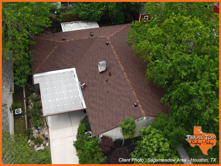Aerial view of a brown shingled roof with white solar panels surrounded by dense green trees and landscaping