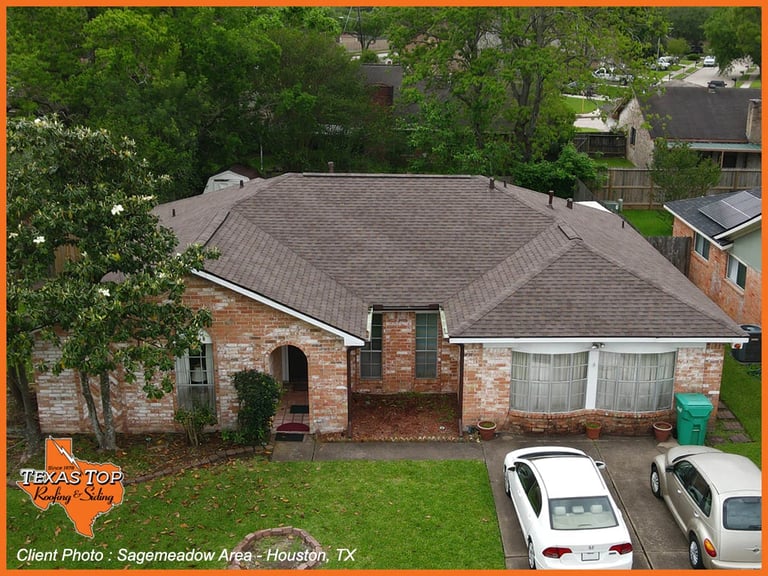 Aerial view of a brick residential home with gray roof, driveway with two parked cars, surrounded by green lawn and trees